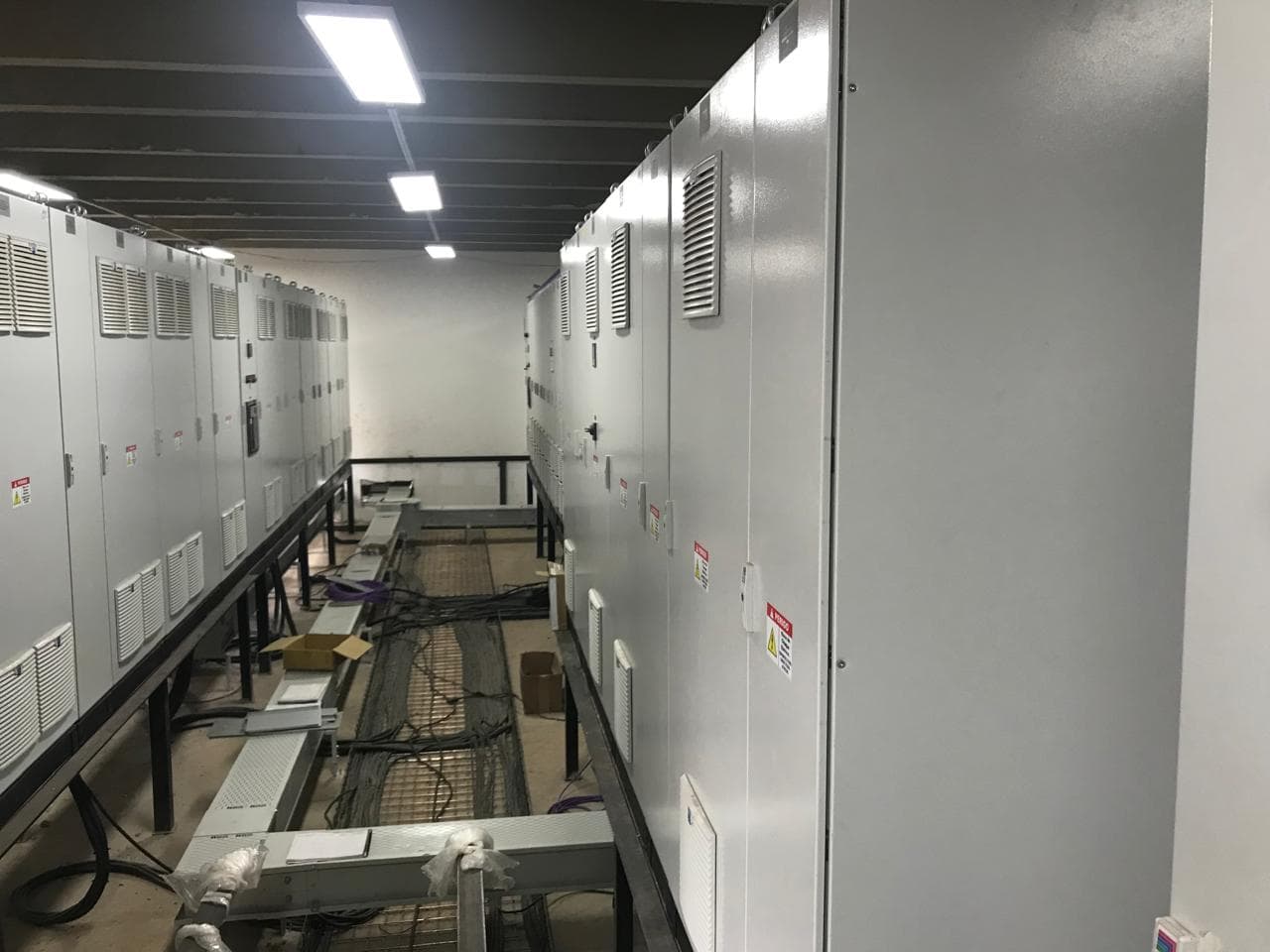 Sala eléctrica con paneles grises ventilados a ambos lados de una bandeja de cables